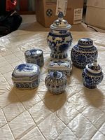 Full set of 7 blue and white porcelain decorative jars of various shapes and sizes, placed on a bedspread with cardboard boxes in background.