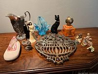 Wide shot of all 14 decorative items arranged on wooden surface showing metal pitcher, blue glass figurine, black cat figurine, metal trivet, ceramic shoe, orange ceramic box with globe lid, and multiple small figurines.