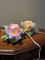 Two porcelain bisque flower-shaped lights, one rose and one iris, shown side-by-side on a wooden surface with white electrical cords attached.