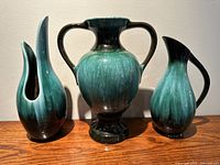 Front view of all three ceramic pieces on a wooden surface showing the urn in center, pitcher on right, and vase on left.