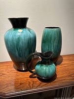 Three ceramic vases arranged on wooden surface showing front view and partial sides. Large vase with wide neck, medium oval vase, small pitcher vase with spout and handle visible.