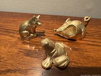 Three brass animal figurines arranged on wooden surface: a detailed fox, an abstract curled tail animal, and a smooth stylized bird.