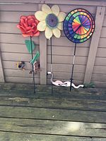 Full view of three pinwheels: red flower, yellow daisy, and multi-colored rainbow pinwheel, with two pink flamingos and a Wiley Coyote chasing Road Runner figure placed on wooden deck by fence.