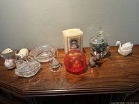 Photo showing nine decorative items arranged on a wooden surface including porcelain boot, glass swan, red vase, candle holder, mouse figurine, and round dishes.