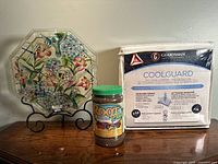 Glass plate with hummingbirds and flowers, ROOT Blast fertilizer jar, and packaged mattress protector displayed together