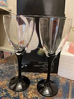 Two clear water goblets with black stems standing next to box featuring floral design and product label.