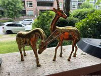 Two gold brass gazelle figurines standing side by side outdoors on a ledge, one with head lowered and one with head raised; background includes a street and buildings.