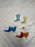 Six miniature shoes and boots displayed on quilted fabric, showing diversity in color and style including blue, yellow, amber, and white with floral designs.