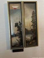 Pair of framed antique pastel landscape paintings hanging on a wall, showing lakeside scenes with trees under muted sky, frames with gold and black trim.