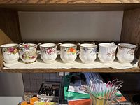 Seven Royal Albert bone china teacups with varied floral patterns arranged in a row on a shelf.