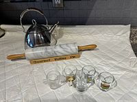 Full view of tea kettle, marble rolling pin on wooden stand, and five shot glasses arranged on a quilted white surface.