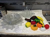 Full lot showing clear pressed glass sunflower patterned platter, basket with handle and multicolored glass fruit figurines arranged on cloth-covered surface.