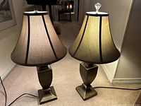 Pair of traditional bronze finish table lamps with beige fabric shades and one having a clear glass finial, placed on carpeted floor in a hallway.
