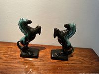 Both ceramic horse bookends shown side by side on wooden surface, rearing pose, black with green mane and tail.