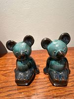 Two bear cub ceramic figurines glazed in green and black, seated on a wooden surface near a beige wall.