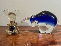 Photo shows two glass animal figurines on a wooden surface: a deep blue and clear glass bull with curved horns, and a smaller mouse/koala with golden ears and blue body details.