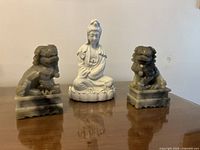 Three statues displayed on wood surface: white female goddess porcelain statue in the middle seated on lotus base, flanked by two gray soapstone Fu Dog statues on pedestal bases