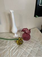 Vintage white Fenton milk glass hobnail 8-finger pedestal vase, cranberry Rossi glass quilted ruffled vase, and vintage glass yellow flower with green stem displayed on a quilted surface.