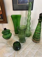 Set of six green glass items including twisted snake bottle, fluted vase, two lidded jars, patterned vase, and gold-accented decanter.