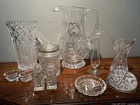 Overview of all crystal glassware items in the set on wooden surface including water jug, jam jar, two small vases, tall vase, salt and pepper shakers, coaster, and paperweight.