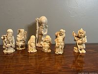 Seven miniature resin Chinese figures arranged standing on wooden surface against a gray wall. Figures vary in detail and size from 3 to 5 inches.