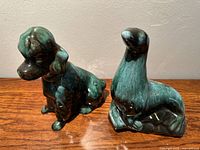 Photo showing both ceramic figurines on wooden surface against neutral wall background. Dog on left, sea lion/seal on right.