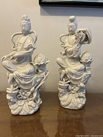Pair of white porcelain Chinese statues seated on lotus flower bases shown side by side on wooden surface.