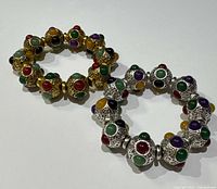Two bracelets side-by-side on a white surface, showing the gold tone bracelet with red, green, black, and purple stones and the silver tone bracelet with similar multicolored stones.
