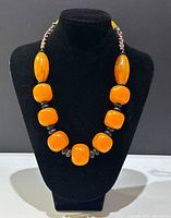 Front view of boho-style chunky beaded necklace on black display stand showing large oval and square simulated butterscotch amber beads with smaller dark accent beads.