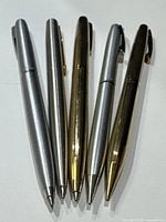 Five collectible pens and pencils arranged side-by-side showing both gold and silver finishes and clip designs.