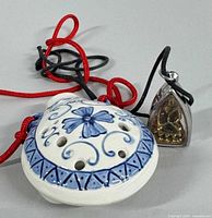 Photo showing the porcelain hand-painted ocarina with blue floral pattern and red cord, alongside the Tibetan brass Buddha pendant in metal case with black cord.