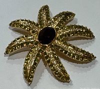 Front view of the vintage Jackie Orr brooch showing the star shape with six arms, each decorated with green rhinestones and centered by a dark oval stone in gold-tone metal.