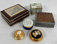 Six vintage collectible trinket boxes arranged on white background including wooden box with sterling silver lid, metal box with flower petals under glass, mother-of-pearl cube box, blue and white floral enamel box, and two gold tone pearl-embellished boxes.