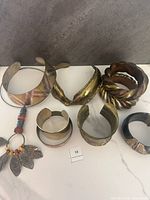 Top-down photo of a collection of nine vintage metal and mixed material bangles and cuffs laid flat on a grey surface showing designs and varied materials including brass, copper, wood, and beads with metal leaf charms.