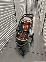 Full view of the Graco jogging stroller showing the three wheels, the orange and charcoal gray seat, and the handlebar, taken in a storage corridor.