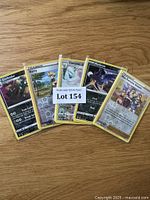 Photo showing a fanned view of five Pokémon cards laid on a wooden surface, including Garbodor, Trainer Barry, Cincinno, Mimikyu, and a Trainer card showing multiple characters. Card borders and details are visible for identification.