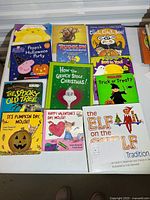 Top-down view of 10 colorful children's picture books arranged in two rows. Titles are seasonal holiday themed including Halloween, Christmas, Thanksgiving, and Valentine's Day. One book has a light-up feature on the cover (Rudolph the Red-Nosed Reindeer).