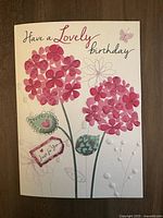 Front close-up of a premium embossed birthday card showing pink floral bouquet, butterflies, and heart-accented leaves with text 'Have a Lovely Birthday'.