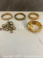 Five vintage gold-tone and mixed-metal bracelets and bangles displayed on a white surface. Includes charm bracelet and varied textured bangles.
