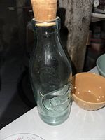 Clear glass bottle with cork stopper, embossed design on the side, placed beside mustard-colored bowl.