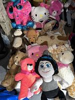 Photo showing multiple stuffed animals including a pink and blue plush, Hello Kitty doll, brown monkey, pink teddy bear, character doll with blue hair, and giraffe plush.