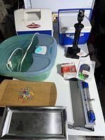 Photo showing miscellaneous household items including a Conair foot spa, two Playmate coolers, a wooden tray, a small metal tray, a black manual hand drill, measuring devices, and a small boxed electronic device.