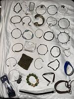 Top-down view of over 30 bracelets and armbands spread on a white cloth, showing a variety of materials including metal, beads, leather, and fabric. Includes spiked bangles, beaded bracelets, open cuffs, and clasps.