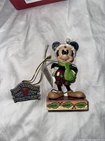 Mickey Mouse figurine standing on a holly decorated base, holding a green scarf, wearing a red and black plaid outfit and blue plaid winter hat.
