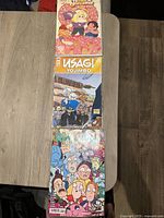 Top-down view of three sealed comic books stacked vertically on a wooden table: Steven Universe 2016 Special, Usagi Yojimbo, and Raanan.