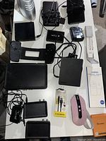 Overview of lot showing miscellaneous small electronics, power adapters, remote control, mouse, and cables arranged on a white surface