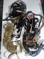 Pile of mixed belts including black leather, striped fabric, faux fur trimmed belts.