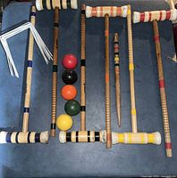 Photo showing six wooden croquet mallets, five colored croquet balls, and a set of white metal wickets on a blue surface.