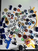 Photo showing scattered Beyblade Burst tops and parts including energy layers, forge discs, performance tips, multiple ripcord launchers in blue and black, and yellow string launchers.