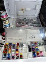 Clear plastic organizer box with many small bobbins of colored thread and cords, plus loose ribbon spools